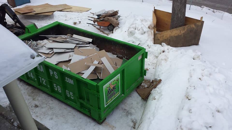 A green dumpster filled with cardboard and debris sits in a snowy outdoor area near a wheelbarrow and wooden enclosure, likely part of a construction site in Long Island, NY.