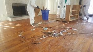 Three individuals in white protective suits cleaning up debris from a room with wooden flooring, a white brick fireplace, and vertical blinds, during a renovation or cleanup project in Long Island, NY.