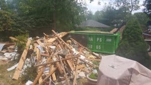 A pile of wooden debris and construction waste is scattered on the grass next to a green dumpster rental in Long Island. The area, surrounded by trees, has a covered object in the foreground. In the background, a house roof peeks through, highlighting the need for efficient junk removal services.