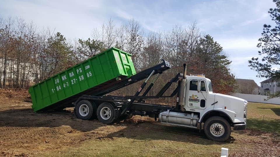 A photo shows a junk removal truck from Long Island, NY. The truck is white with a green dumpster on a tilting platform, ready to unload. The background includes a grassy yard with trees and houses under a clear sky. The company's contact information is visible on the side of the dumpster.