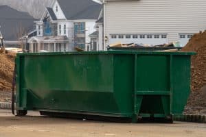 A large green dumpster with visible wear placed outdoors near a white-walled building with multiple doors, under daylight in Long Island, NY.