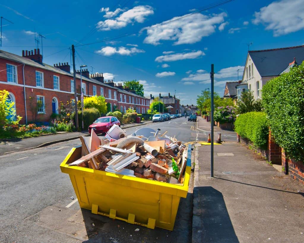 A large yellow dumpster filled with construction debris sits on a residential street lined with brick houses and parked cars under a partly cloudy sky in Long Island, NY.