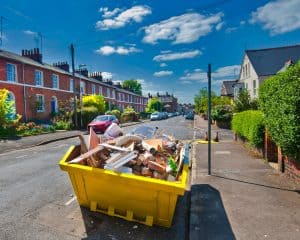 A large yellow dumpster filled with construction debris sits on a residential street lined with brick houses and parked cars under a partly cloudy sky in Long Island, NY.