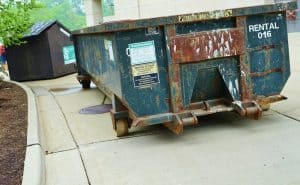 A large rusted metal dumpster labeled 'RENTAL 016' with warning signs, placed on a concrete surface near a smaller black dumpster in an outdoor area in Long Island, NY.