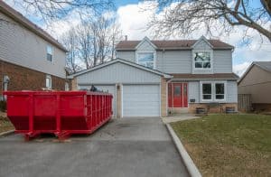 A two-story gray house with a red front door and white garage, featuring a large red dumpster in the driveway, possibly for renovation or cleanup, in Long Island, NY during late fall or winter.