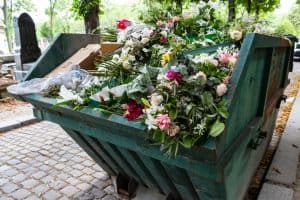 A dumpster filled with discarded flower arrangements that were left behind on graves from visitors at the Père Lachaise Cemetery, Paris, France on a cobblestone road.
