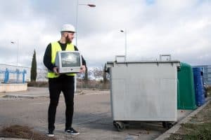 A person in a white hard hat and yellow safety vest holding an old CRT television next to a large gray dumpster on a roadside in Long Island, NY, with trees, streetlights, and recycling bins in the background.