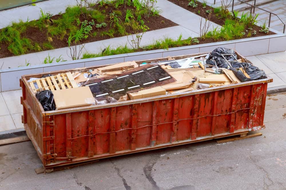 A rusted red dumpster filled with construction debris, including wood planks, cardboard, and black trash bags, sits on a paved lot in Long Island, NY. In the background, there's a landscaped area with fresh dirt, new plants, and a concrete path.